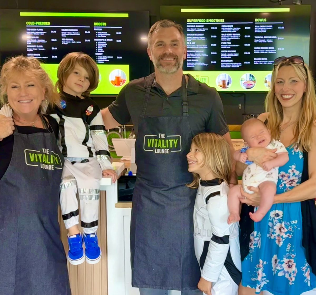 The Vitality Lounge family in matching aprons behind the counter, under the menu boards